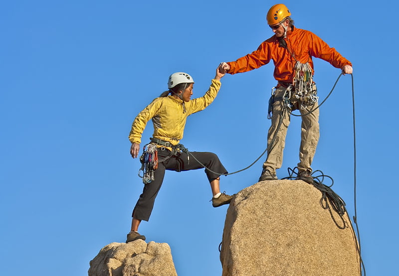 Un guide aide une femme à monter au sommet d'un rocher en hauteur, symbole des bienfaits de la thérapie personnelle | thérapie et coaching - en ligne Sutton, Brome-Missisquoi, Montérégie | Liance