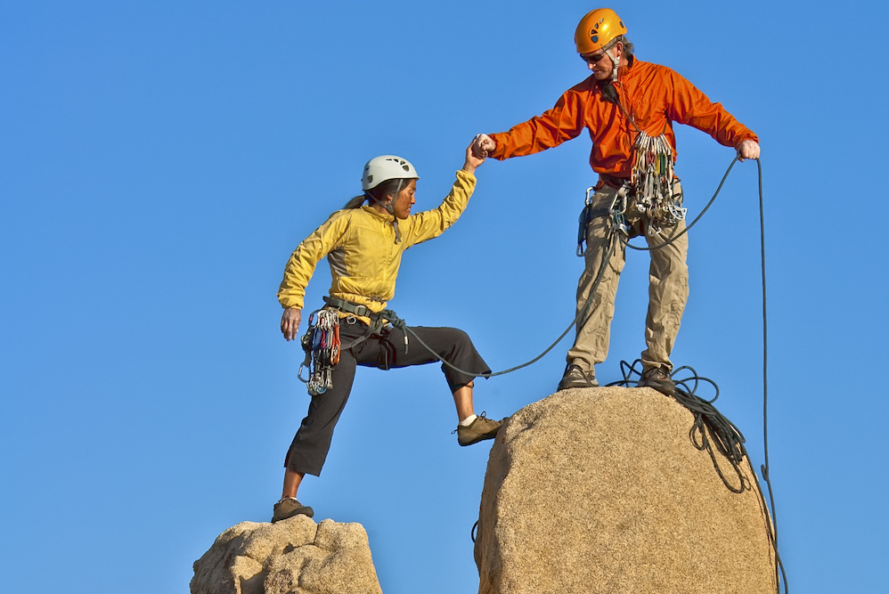 Un guide aide une femme à monter au sommet d'un rocher en hauteur, symbole des bienfaits de la thérapie personnelle | thérapie et coaching - en ligne Sutton, Brome-Missisquoi, Montérégie | Liance