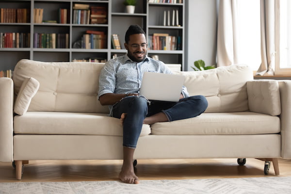 Homme souriant assis sur son canapé avec son ordinateur sur les genoux | thérapie en ligne - Sutton, Québec, Canada, France, Belgique, Suisse | Liance